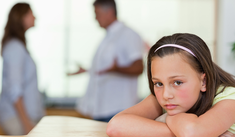 A young girl with a pink headband resting her chin on her arms, with two adults talking behind her