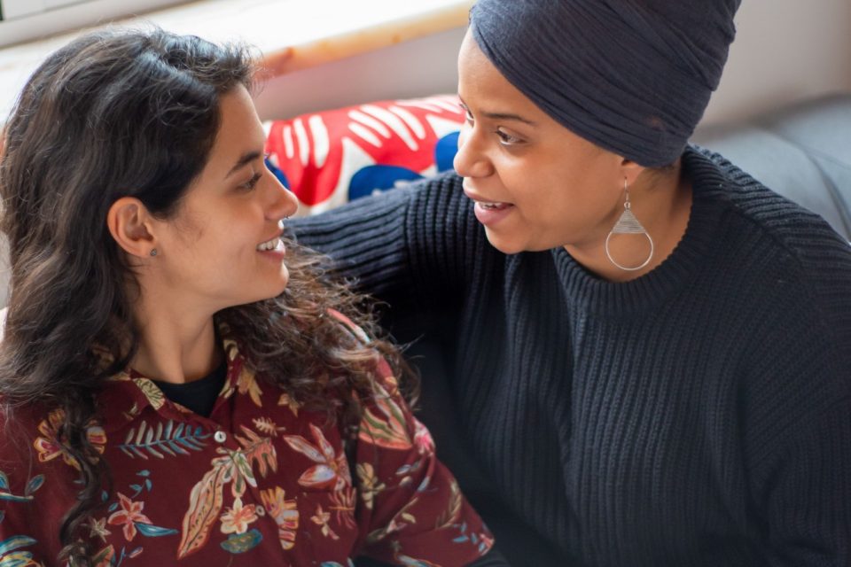 Two women talking and smiling on a couch, one holding a clipboard