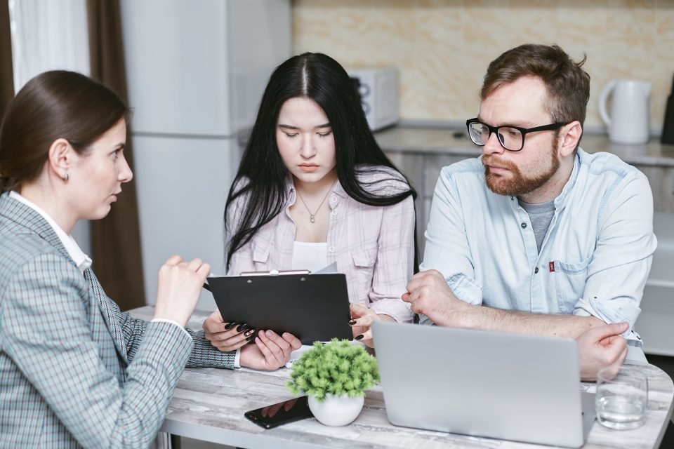 Three people at a table talking over a clipboard and laptop