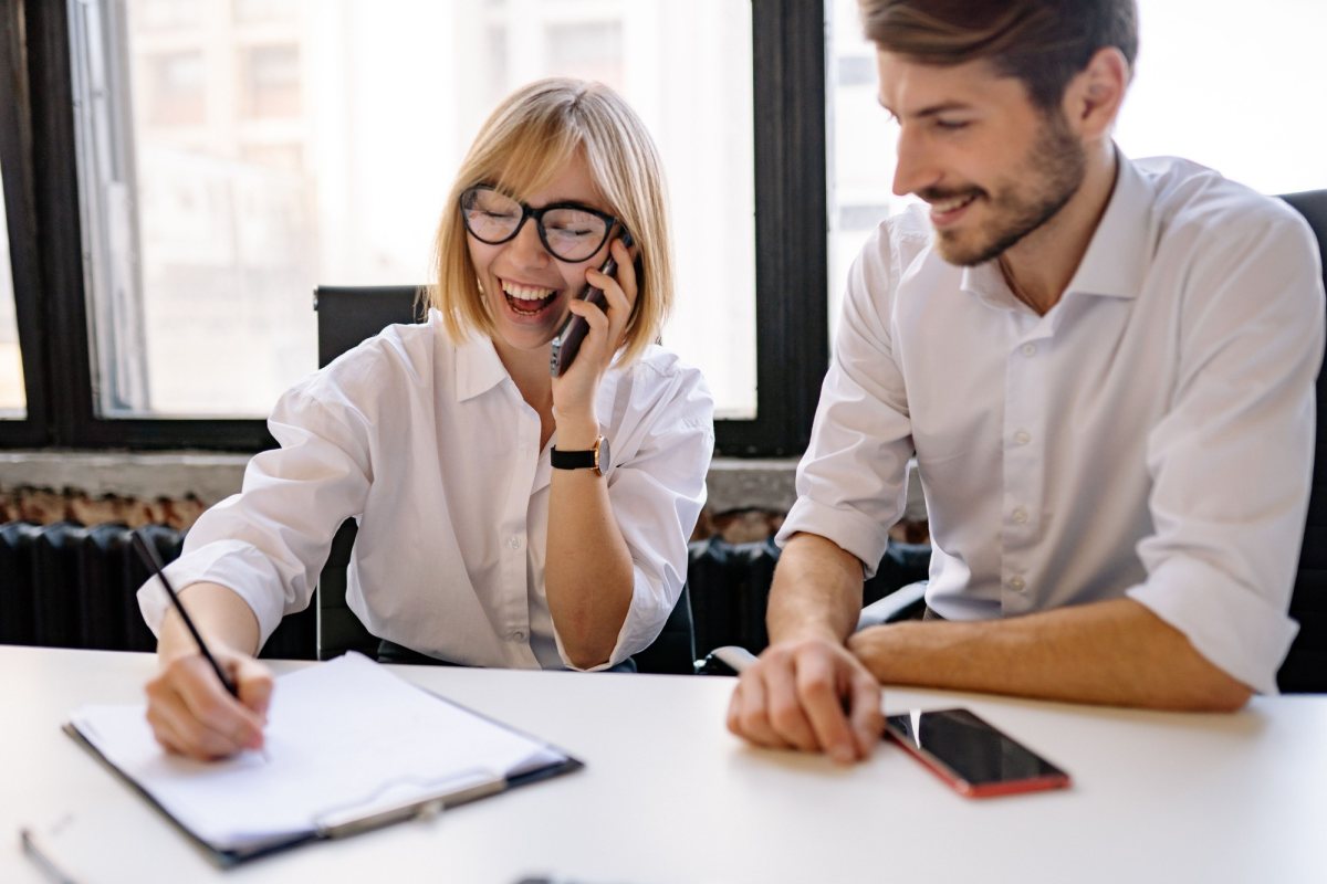 A woman laughing on a phone call while taking notes, with a colleague smiling beside her